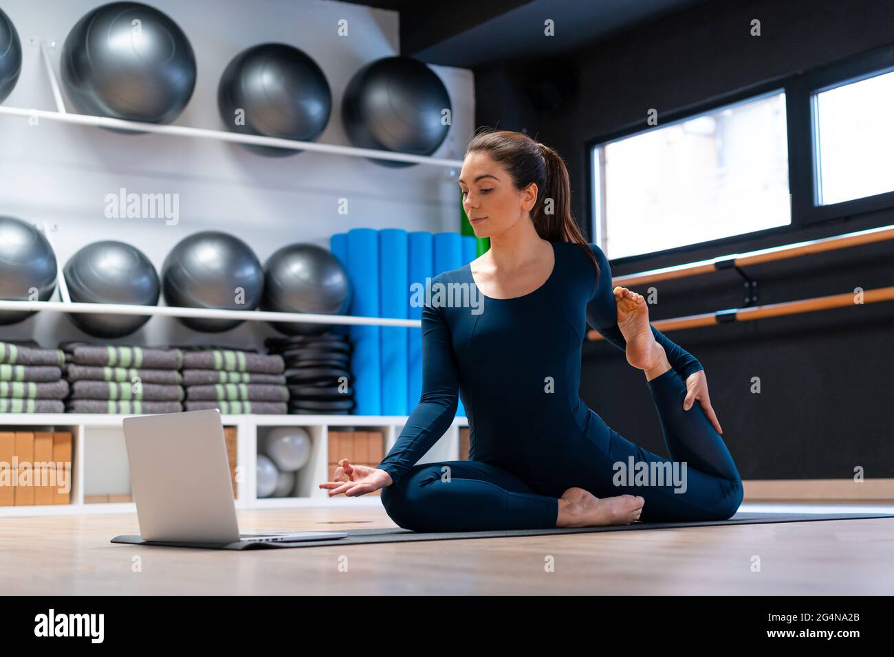 Ganzer Körper der flexiblen jungen weiblichen Yogalehrerin, die eine einbeinige King Pigeon Variation mit Mudra-Geste vor dem Laptop-Bildschirm während onl Stockfoto