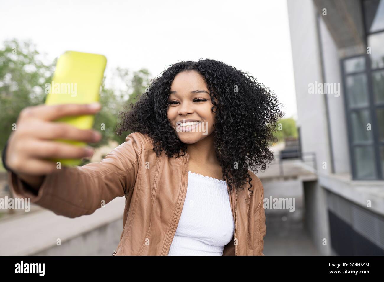 Optimistische afroamerikanische Frau mit lockigen Haaren, die auf dem Smartphone Selbstporträt aufnimmt, während sie auf der Straße in der Nähe des Gebäudes auf unscharfem Hintergrund steht Stockfoto