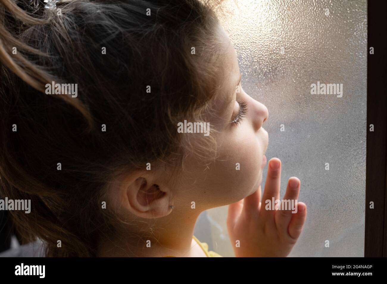 Seitenansicht Kopfbild des neugierigen niedlichen kleinen Mädchens, das neben einem durchsichtigen Glasfenster steht und nachdenklich davonschaut Stockfoto