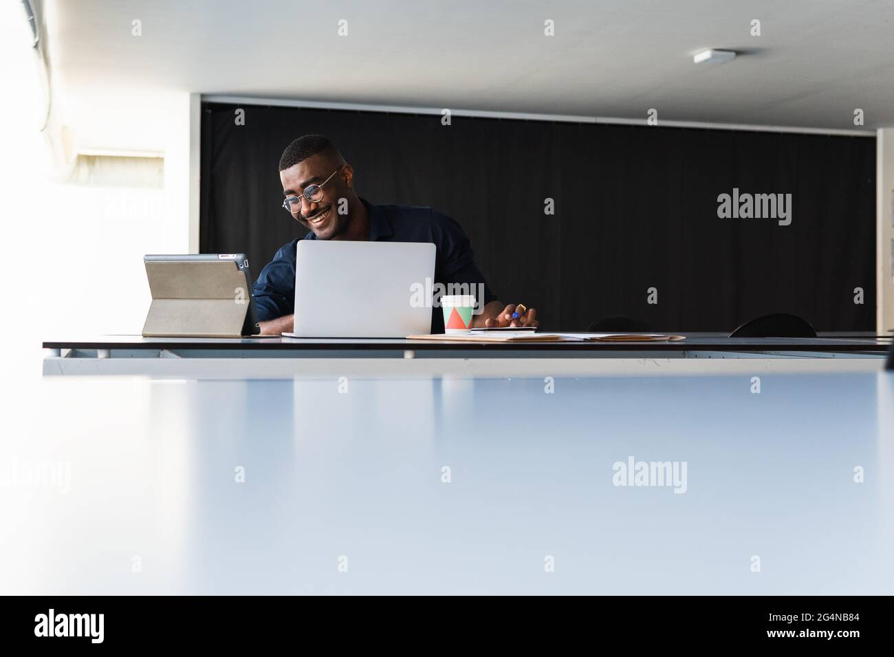 Positiver afroamerikanischer Erwachsener, männlich in formeller Kleidung und Brillen, die am Tisch sitzen, während er einen Videoanruf auf dem Tablet in der Nähe des Netbooks führt und Wi dokumentiert Stockfoto