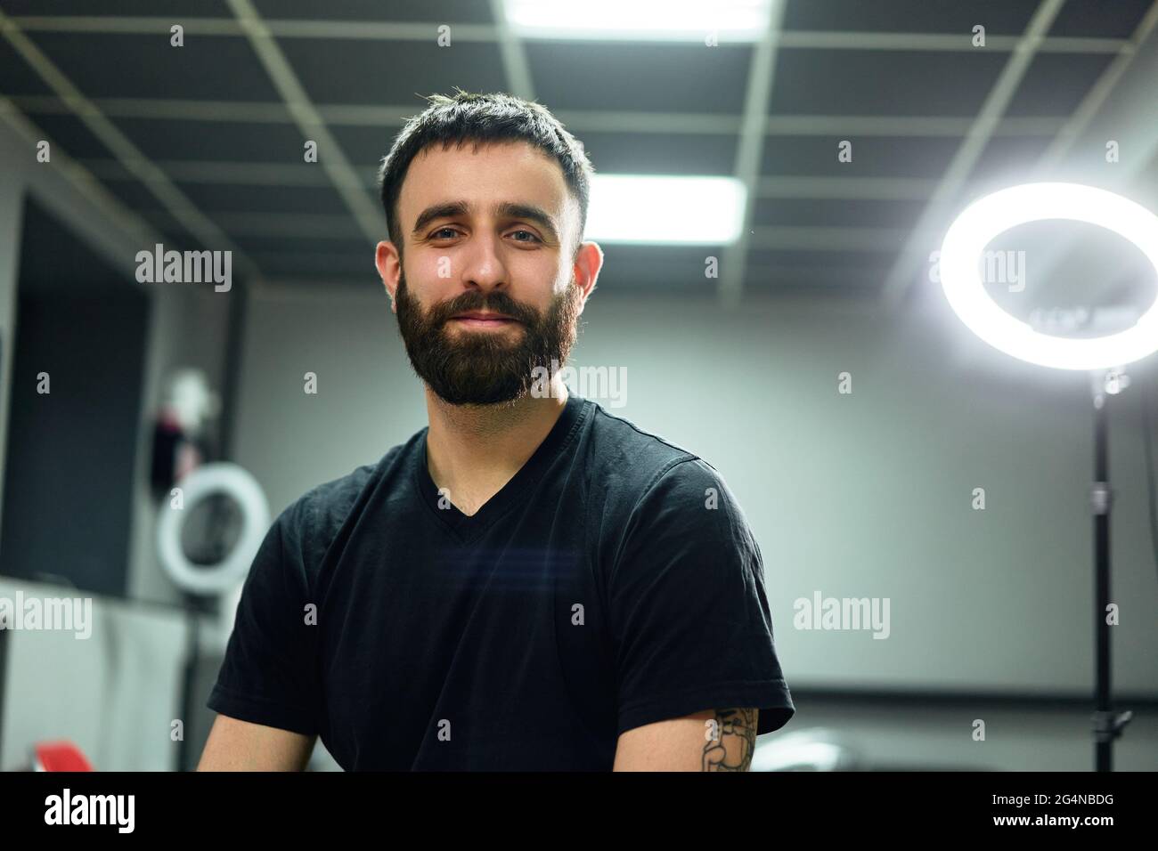 Glücklicher, bärtiger, erwachsener Tätowierer in legerer Kleidung, der auf einem Hocker neben einer Ringlampe und Instrumenten sitzt, während er im hellen Tattoo-Salon die Kamera anschaut Stockfoto