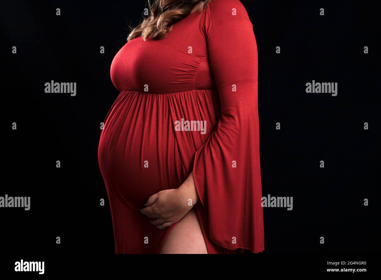 Nicht erkennbare Schwangere, die ein langes rotes Kleid trägt, umarmt ihren großen Bauch, während sie erwartet Stockfoto