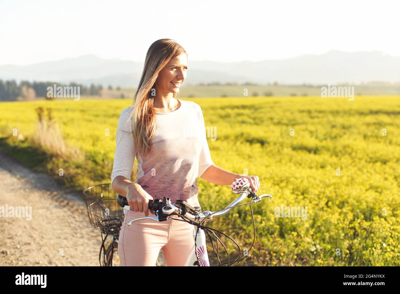 Junge Frau, die neben ihrem Fahrrad über staubige Landstraße steht, starke nachmittägliche Sonnenbeleuchtung im Hintergrund leuchtet auf gelbem Blumenfeld Stockfoto
