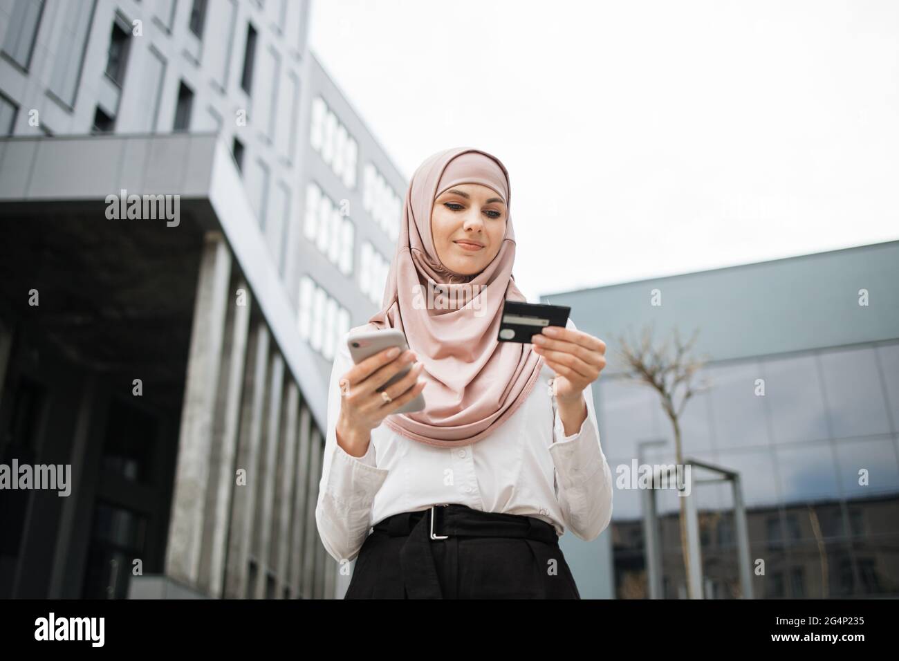 Schöne muslimische Frau in formeller Kleidung und Hijab tun Online-Shopping  mit Kreditkarte und Smartphone. Junge Dame, die in der Nähe des  Bürogebäudes steht und die einfache Bezahlung genießt Stockfotografie -  Alamy