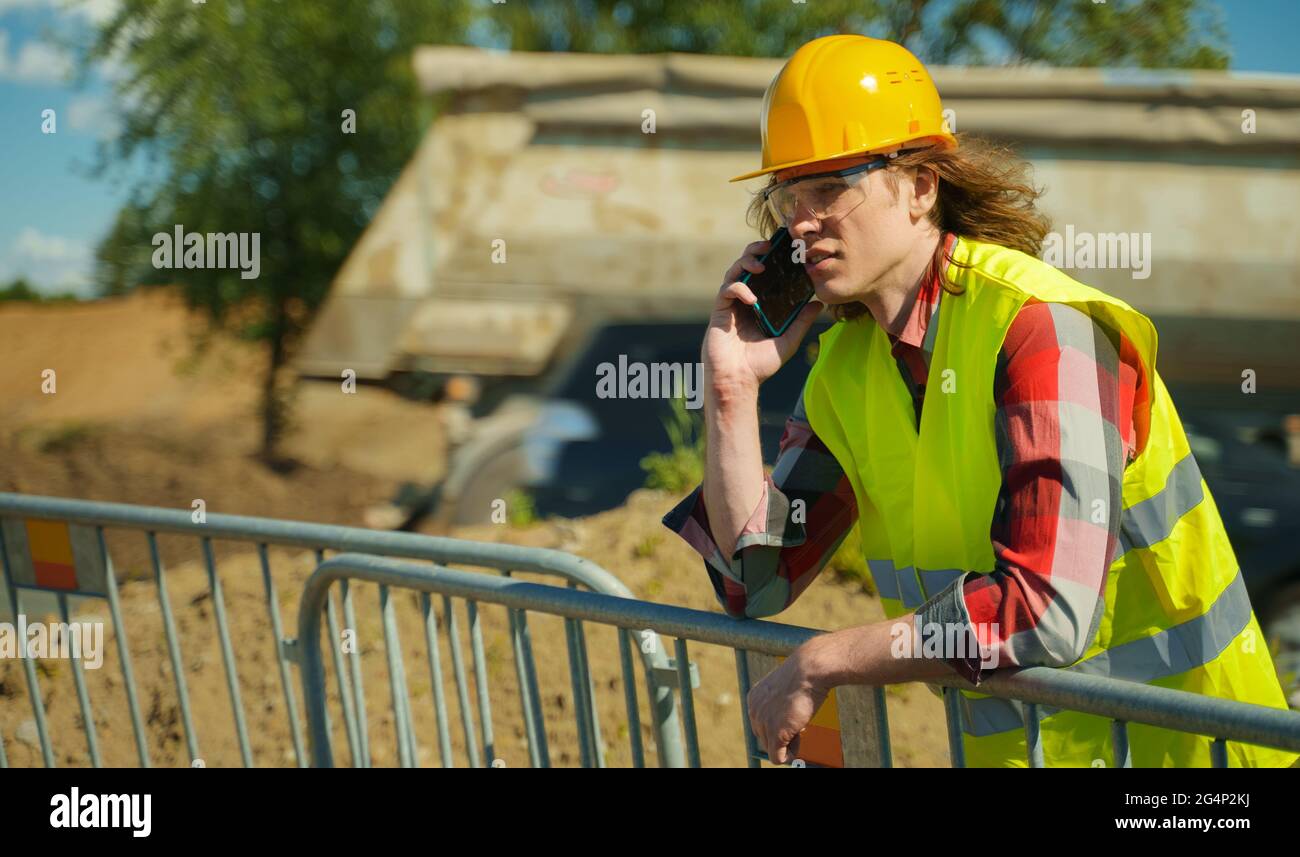 Männlicher Arbeiter in Hardhut und hoher Visusjacke, der mit dem Smartphone spricht. Stockfoto