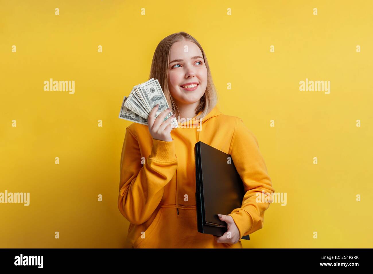 Porträt einer jungen, aufregend lächelnden, hübschen Frau, die einen Stapel Geldscheine und einen Laptop in der Hand hält. Teenager Mädchen träumen von Geld, Einkommen online Stockfoto