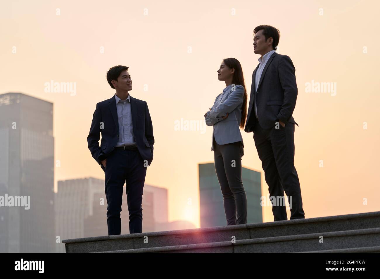 Team von asiatischen Unternehmensleitern, die auf einer Plattform stehen und sich mit Gesprächen unterhalten, die in der Abenddämmerung mit Stadthintergrund unterhalten Stockfoto
