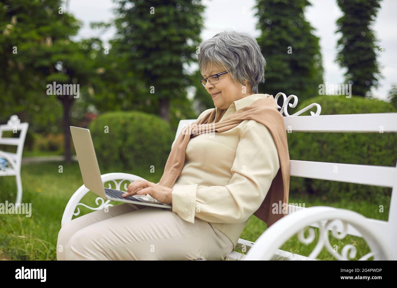 Alte Dame, die einen Laptop benutzt und sich im Park ausruht, während sie auf der Bank sitzt Stockfoto