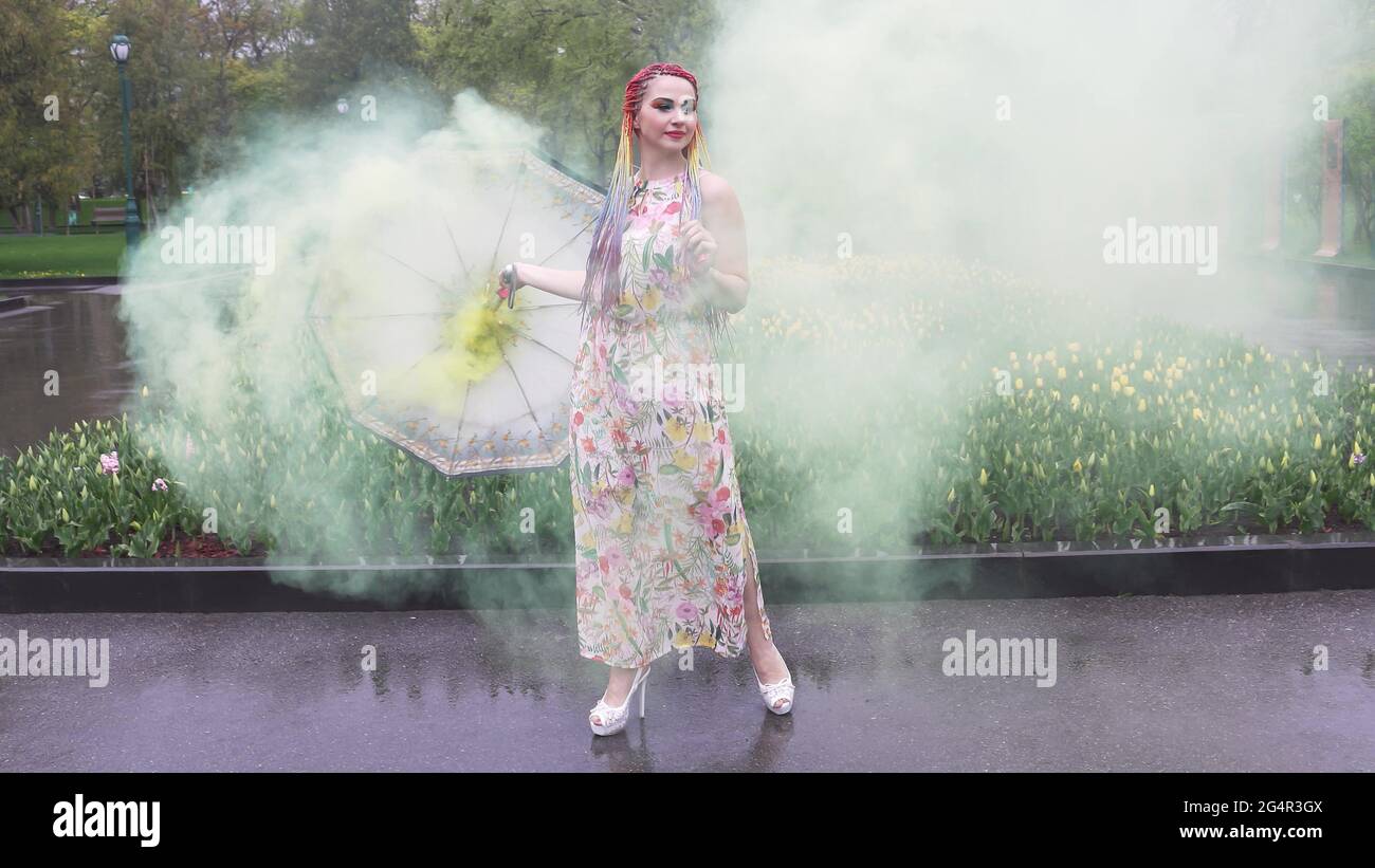 Entzückendes Mädchen in einem Kleid mit einem Blumendruck mit Regenbogen Zöpfen und Make-up. Tanzt mit einem Regenschirm, der sich in gelbem Rauch vor der Kulisse von FL Stockfoto