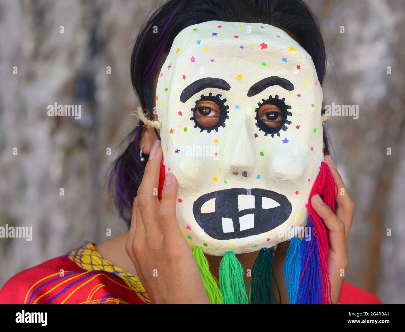Der junge mexikanische Künstler (Maskenmacher, Maskenmaler) zeigt eine seiner traditionellen kulturellen folkloristischen Masken und trägt sie über einer schwarzen chirurgischen Gesichtsmaske. Stockfoto