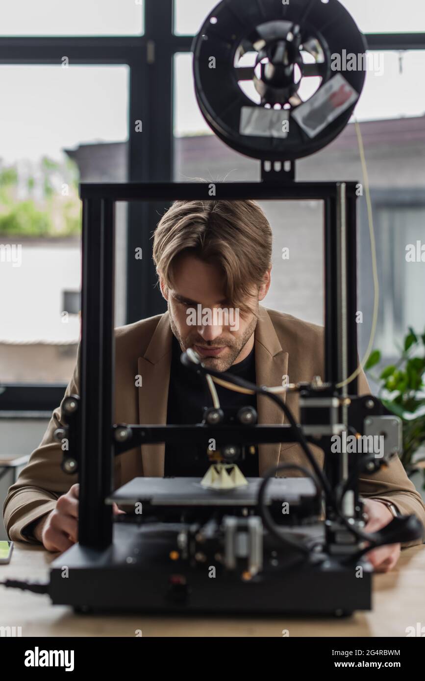 Fokussierter junger Designer, der sich mit dem 3D-Drucker beschäftigt, der im modernen Büro ein Modell herstellt Stockfoto