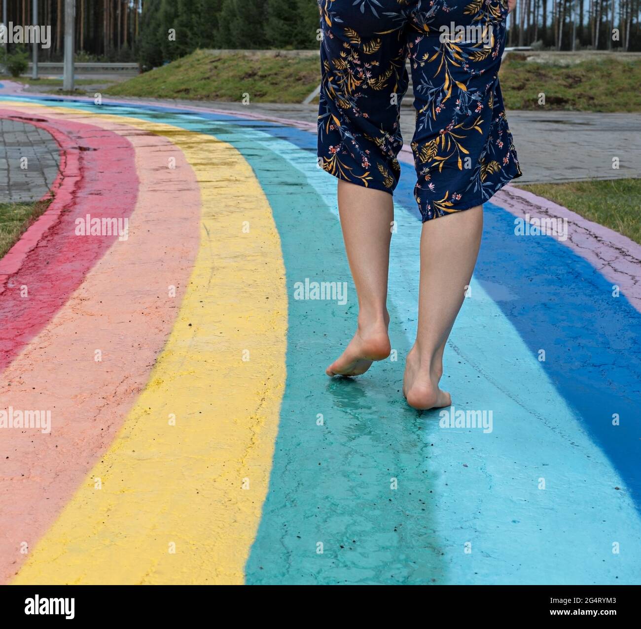 Weibliche Beine schließen sich barfuß auf nassem Asphalt gezogenem Regenbogen an, der zwischen Pfützen, Platz, Liebe, Freiheit und Freundschaft führt Stockfoto