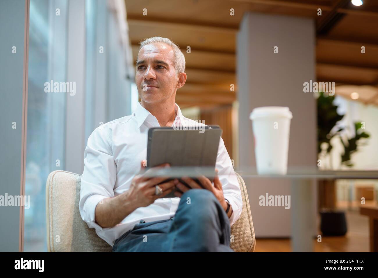 Porträt eines gutaussehenden Geschäftsmanns, der im Café sitzt und ein digitales Tablet verwendet, während er durch das Fenster blickt und eine horizontale Aufnahme durchdenkt Stockfoto