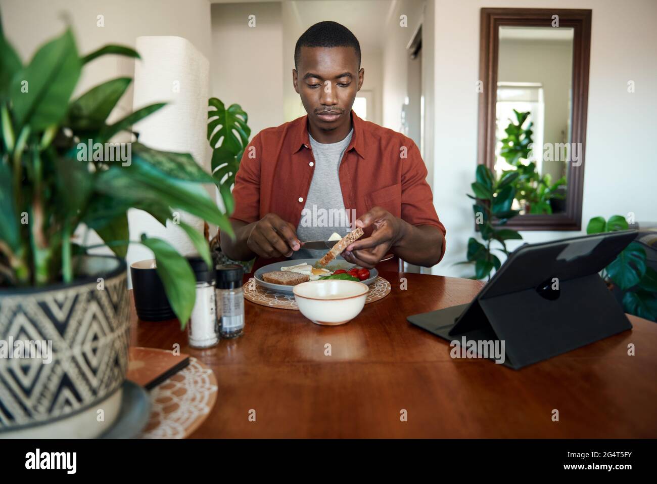 Junger afrikanischer Mann, der eine Tablette benutzt und Frühstück isst Stockfoto