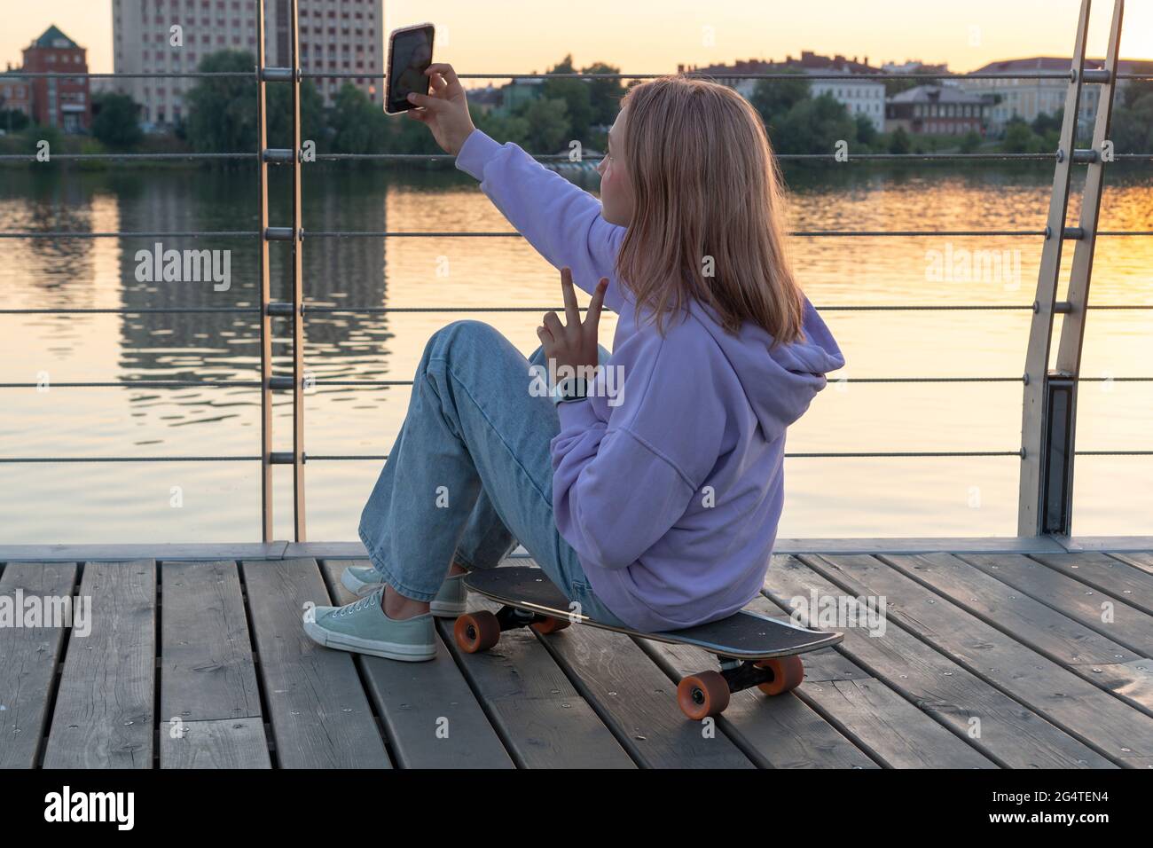 Stilvolles Teenager-Mädchen in einer lila Kapuzenjeans mit rosa Haaren sitzt auf einem Longboard mit einem Telefon in der Hand. GEN Z-STILTRENDS. Stockfoto