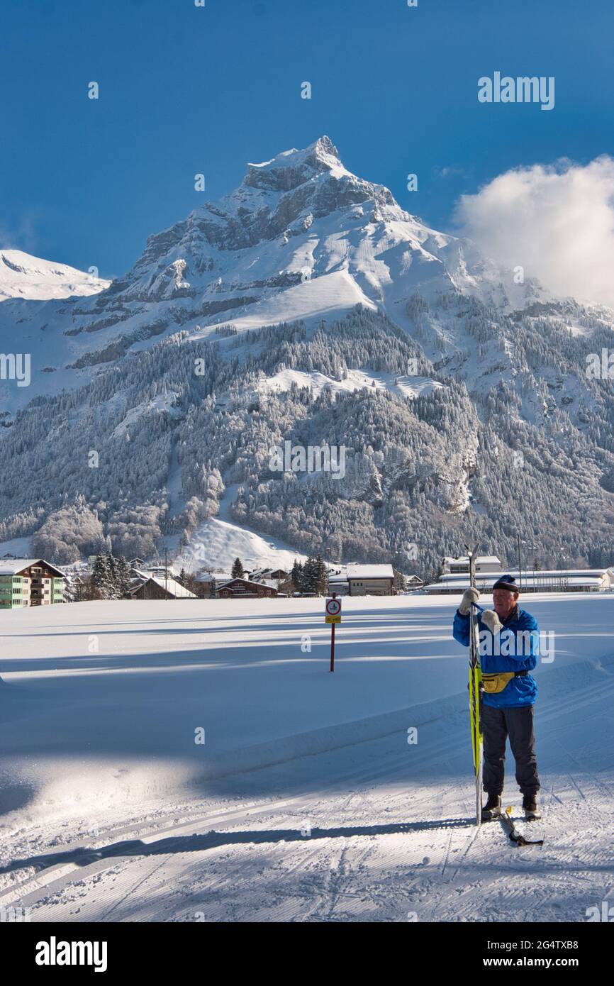 Eine Landschaft von einem sonnenbeschienenen schneebedeckten Feld mit Bergkulisse bei Engelberg, Kanton Obwalden, Zentralschweiz, macht sich ein langlaufiger Skifahrer bereit Stockfoto
