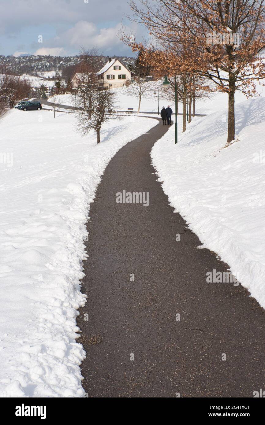 Ein schneefreiter Weg durch einen Park, der sich in die Ferne schlängelt, wobei ein Paar auf beiden Seiten in Oberdorf, Schweiz, mit Schnee zu Fuß unterwegs ist Stockfoto