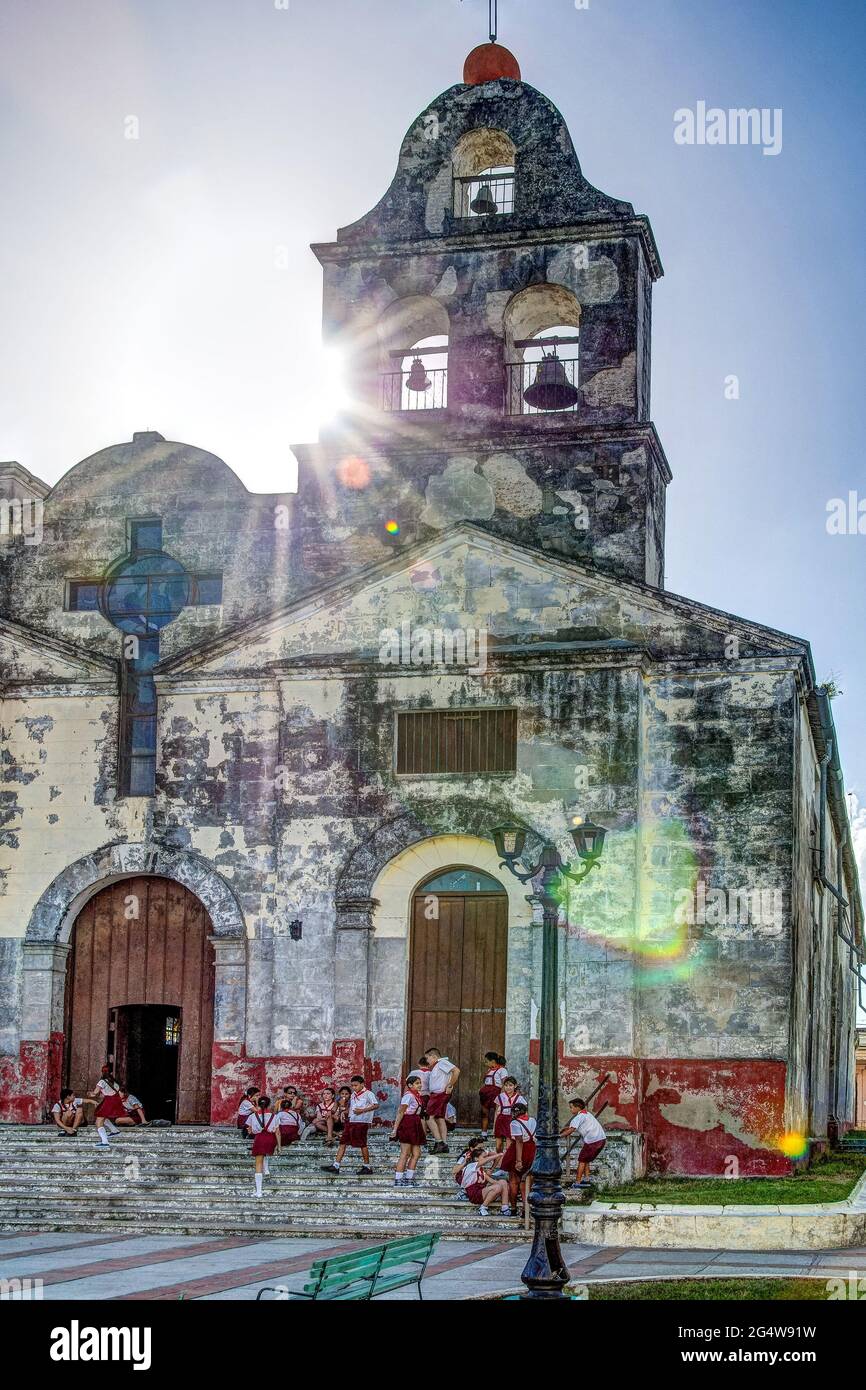 Kubanische Pioniere vor einer alten katholischen Kirche an einem sonnigen Tag mit drei Glocken und Kreuz auf dem Dach, Schulkinder hängen auf Treppen vor der Th Stockfoto