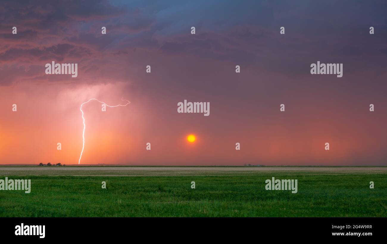Der Blitz trifft bei Sonnenuntergang ein, wobei die Sonne stark gefiltert durch den in der Ferne fallenden Regen gesehen wird. Dunkle Sturmwolken sind über uns zu sehen. Stockfoto