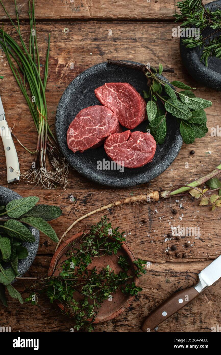 3 rohe Filet-Mignon-Steaks in grauem Steinteller auf rustikalem Holztisch mit Steakmessern und frischen grünen Kräutern Draufsicht Stockfoto