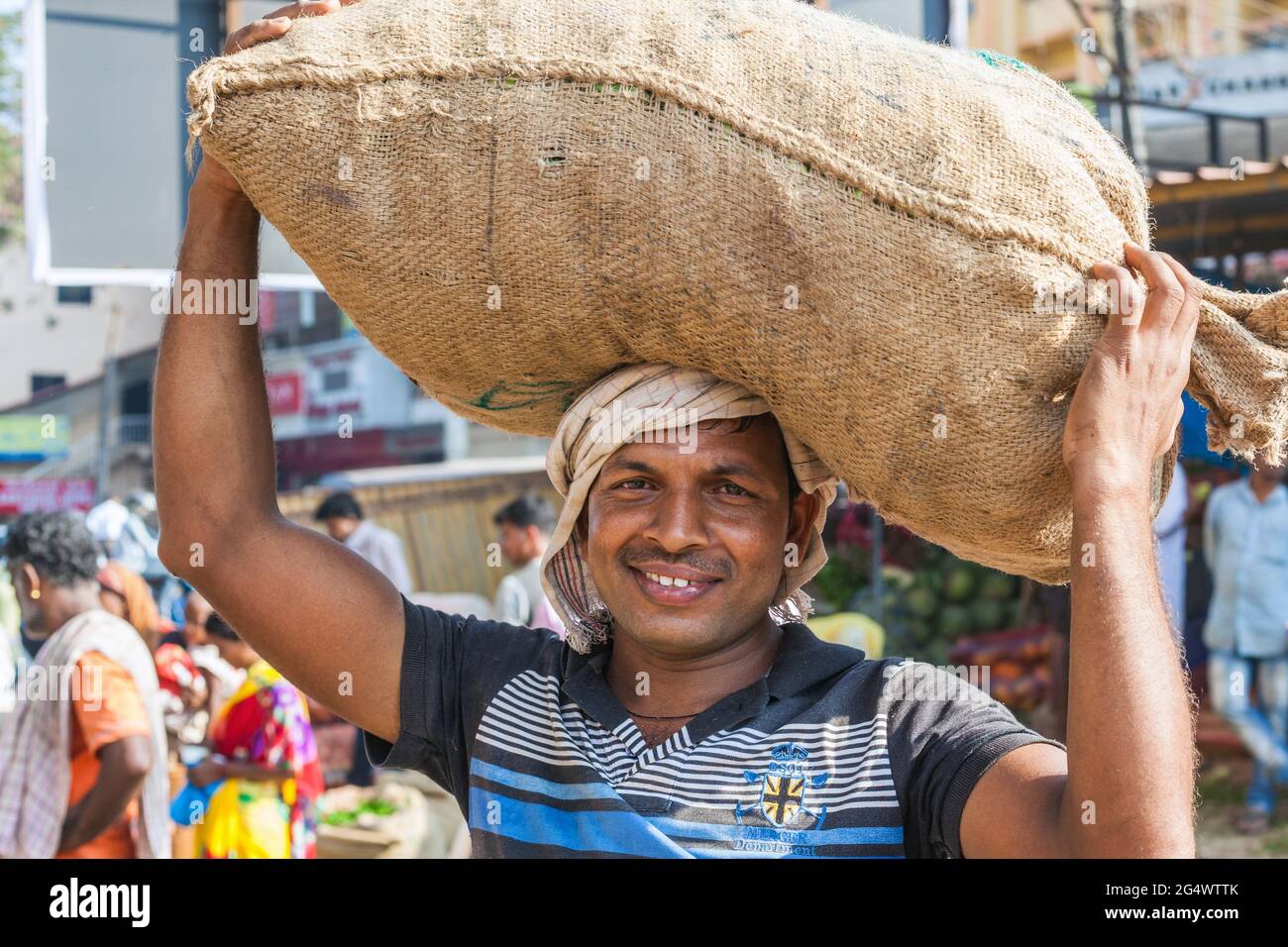 Lächelnder indischer Portier, der einen schweren Sack von Produkten auf dem Kopf trägt, Udupi, Karnataka, Indien Stockfoto