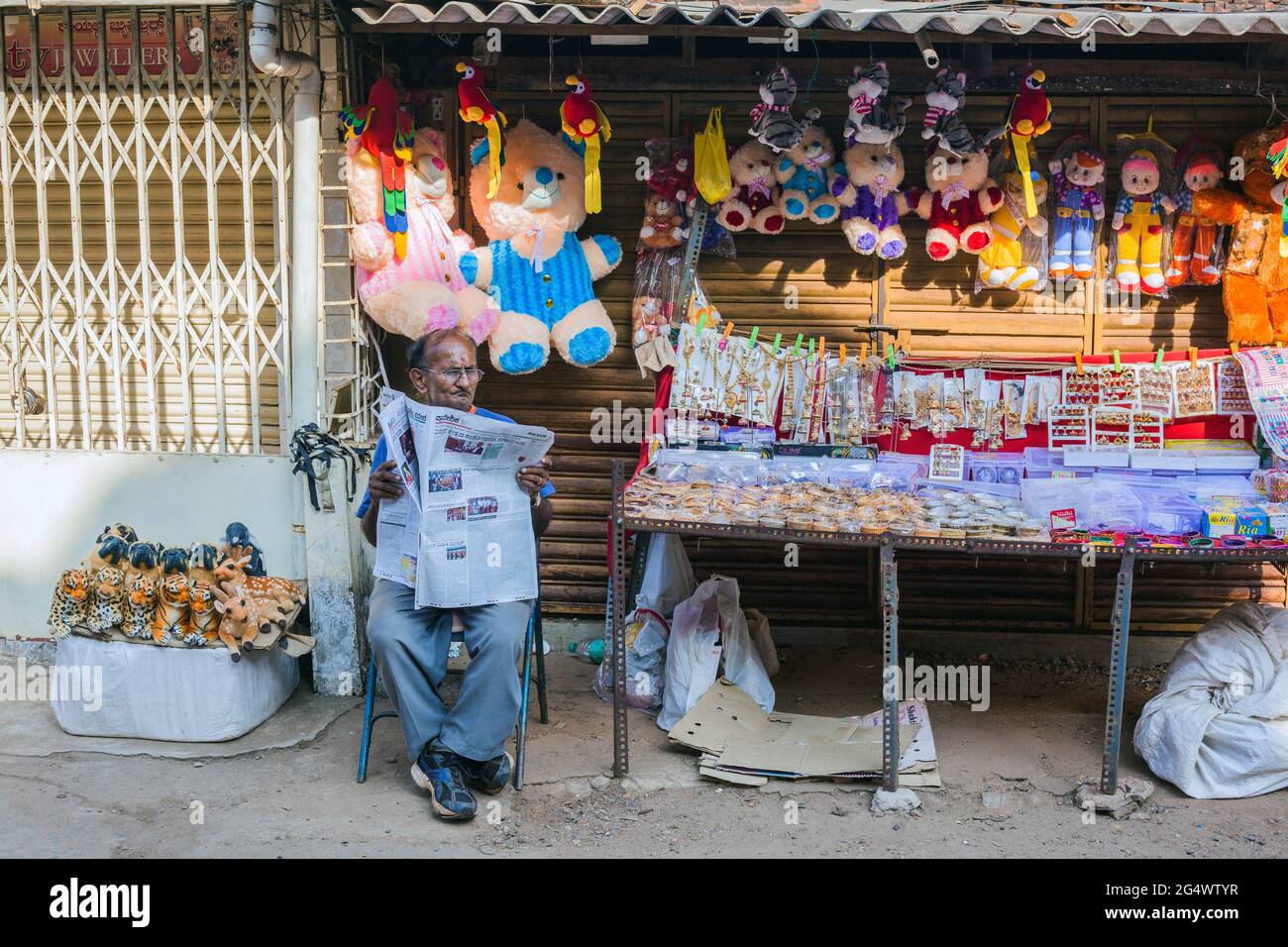 Der kahlen indische Mann sitzt beim Lesen der Zeitung, während er an einem Straßenstand Kuscheltiere und Goldschmuck verkauft, Udupi, Karnataka, Indien Stockfoto
