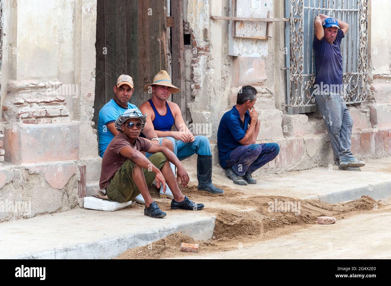 Kubanische Bauarbeiter sitzen gemächlich vor einem alten, teilweise beschädigten Gebäude mit etwas Bausand vorne. Stockfoto
