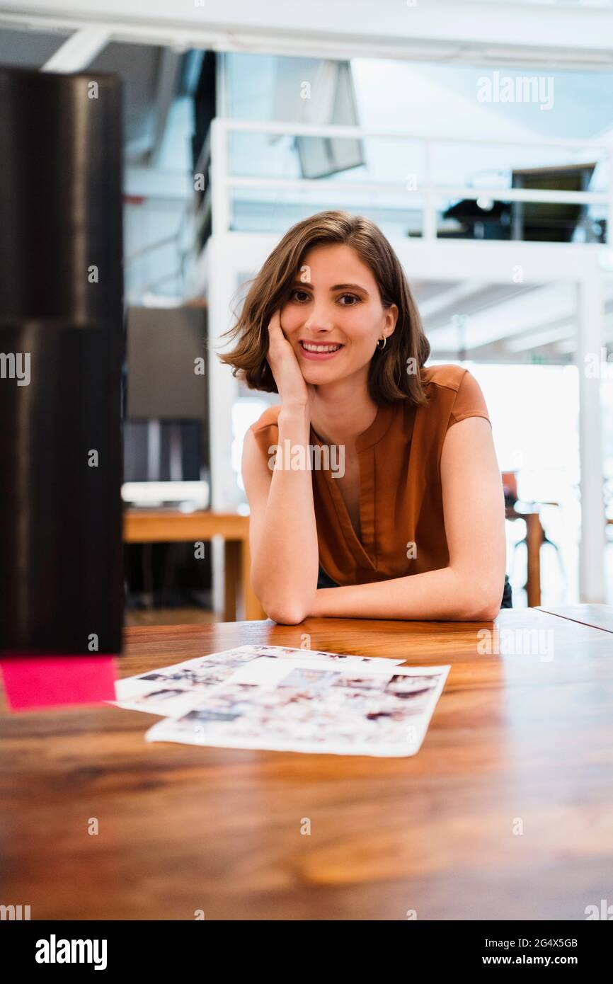 Lächelnde junge professionelle Frau, die im kreativen Büro mit der Hand auf dem Kinn sitzt Stockfoto