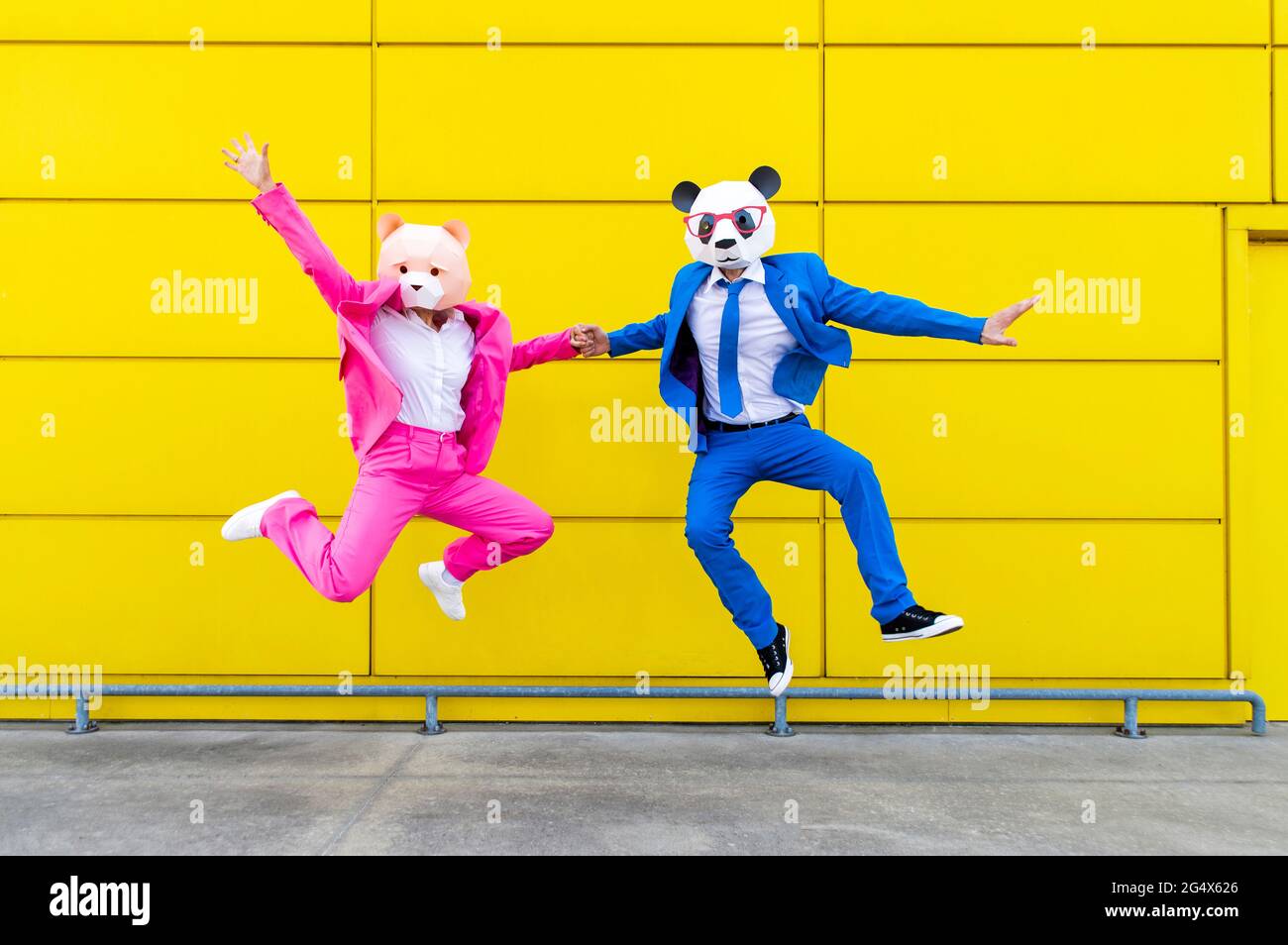 Mann und Frau tragen lebhafte Anzüge und tragen Masken, die die Hände halten, während sie gegen die gelbe Wand springen Stockfoto