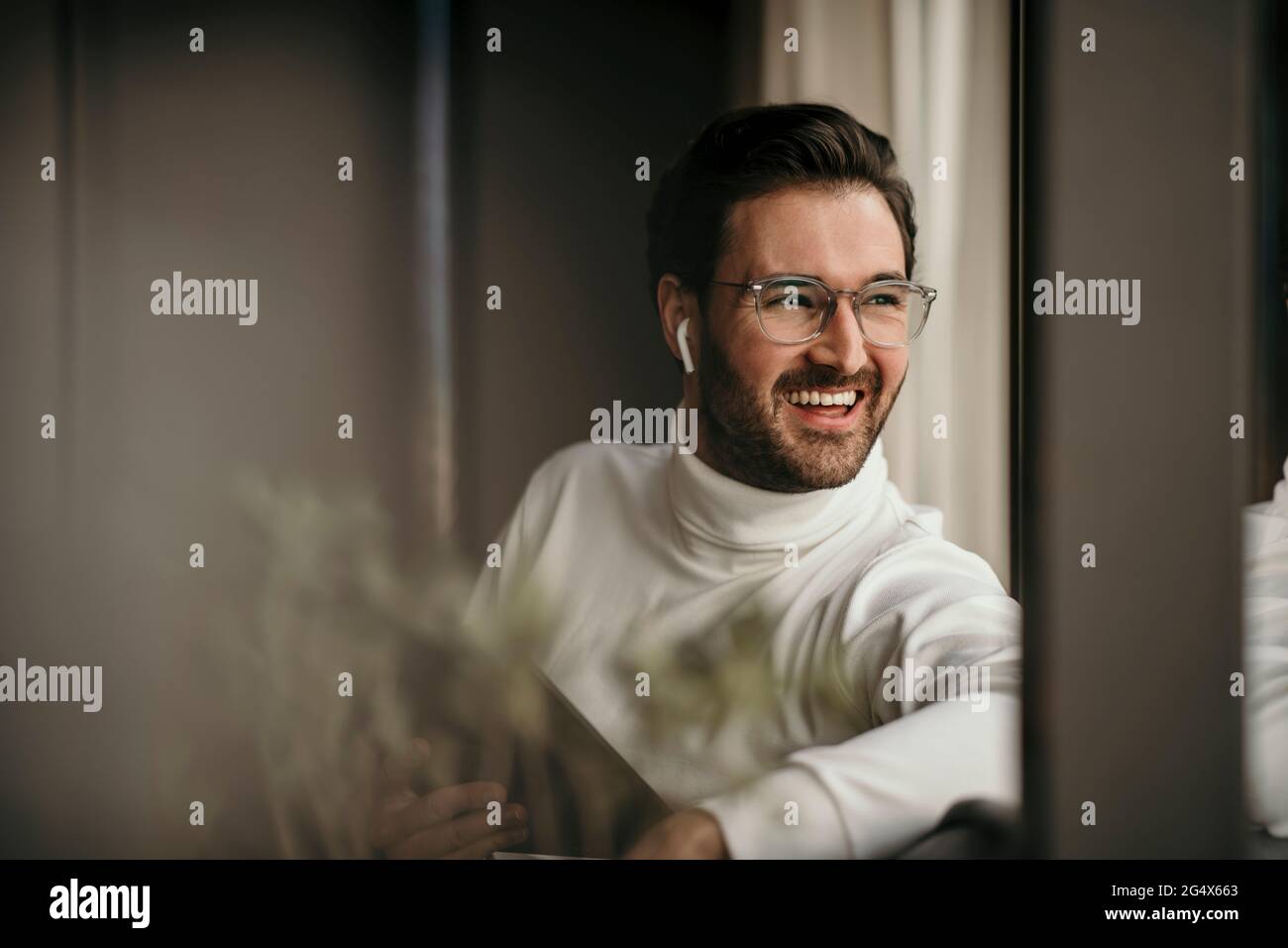 Fröhlicher Geschäftsmann hält ein digitales Tablet, während er zu Hause durch das Fenster schaut Stockfoto