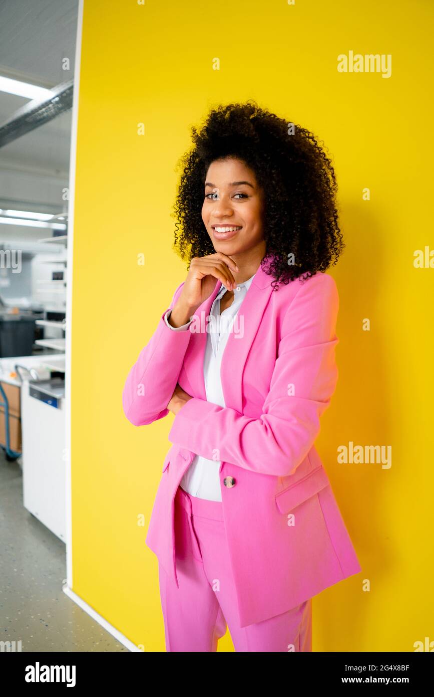Lächelnde Geschäftsfrau mit der Hand auf dem Kinn, die vor der gelben Wand steht Stockfoto