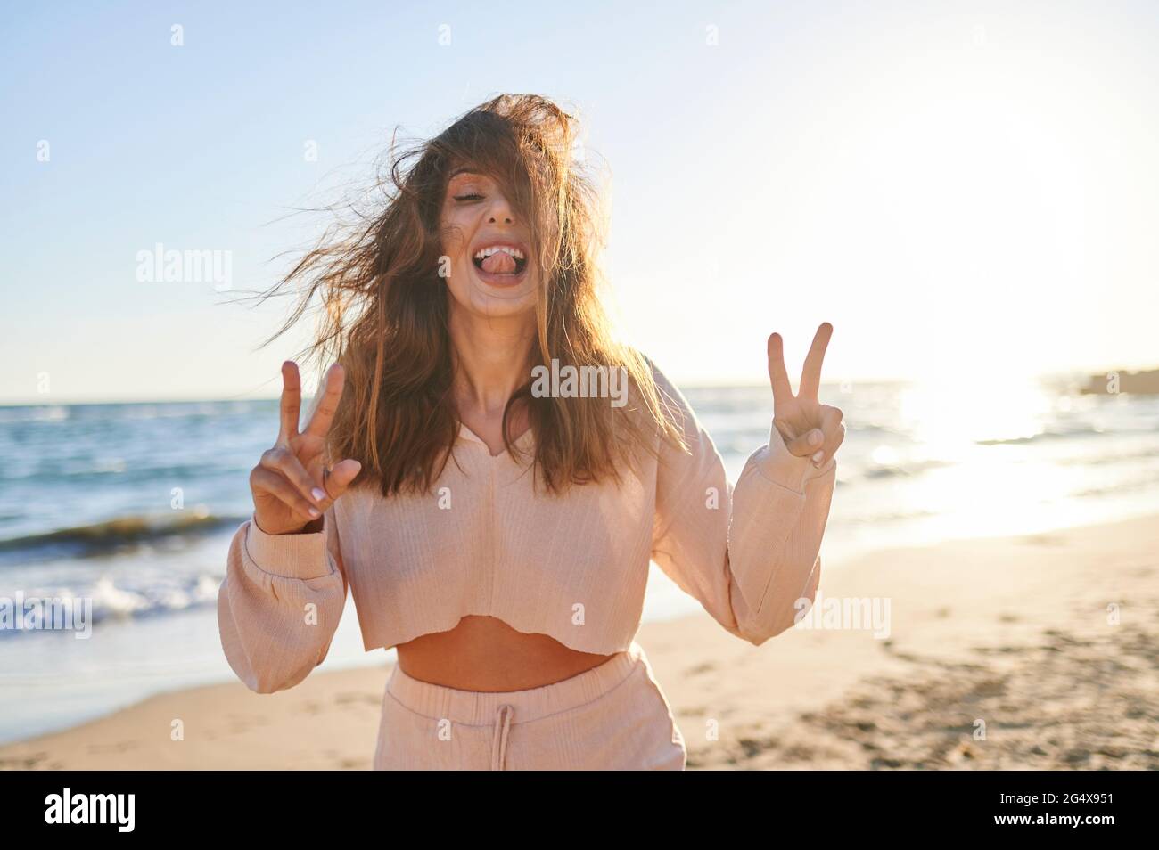Fröhliche Frau mit Friedenszeichen, während sie am Strand die Zunge herausstreckte Stockfoto