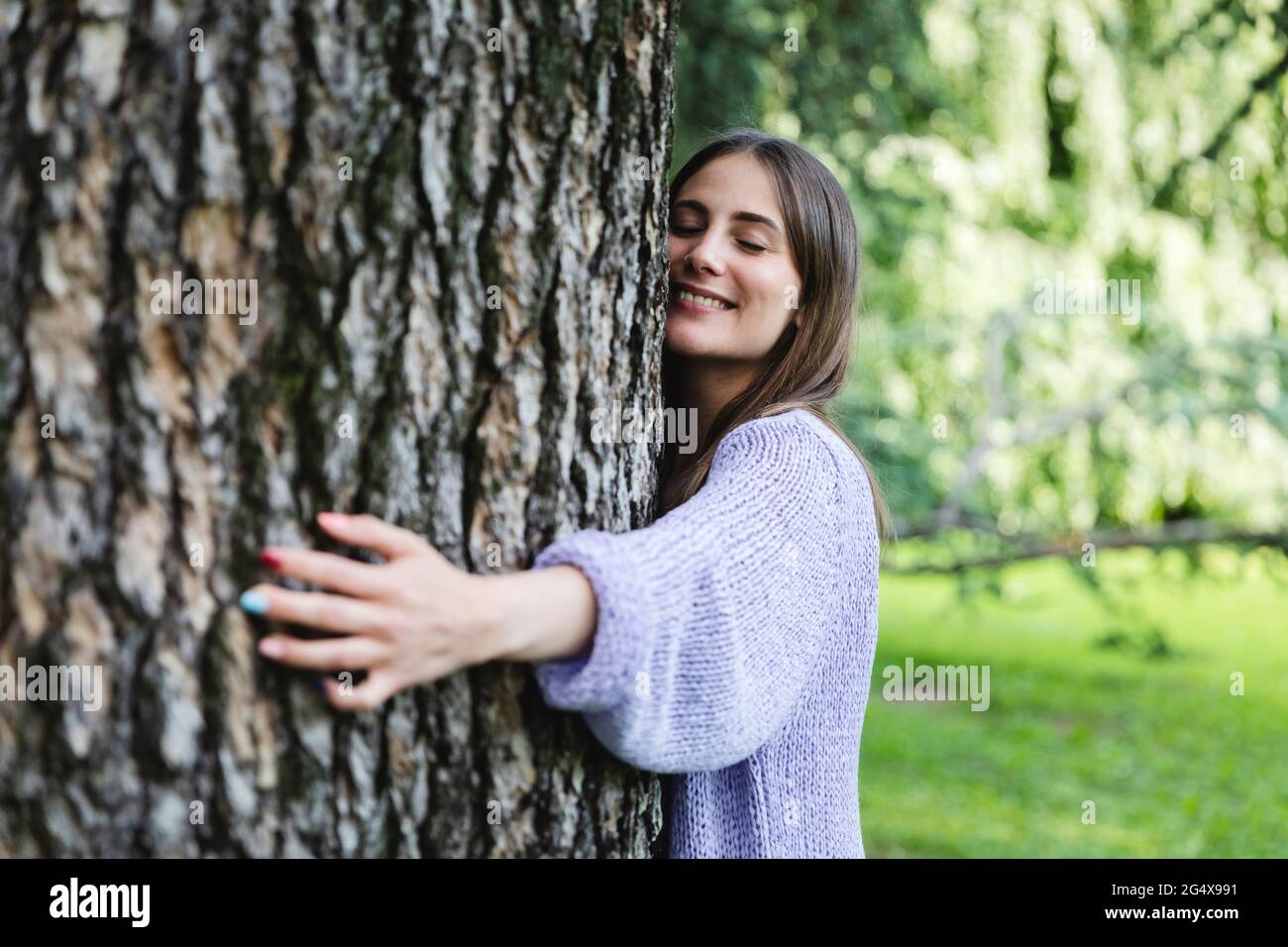 Lächelnde Frau mit geschlossenen Augen umarmenden Baum im öffentlichen Park Stockfoto