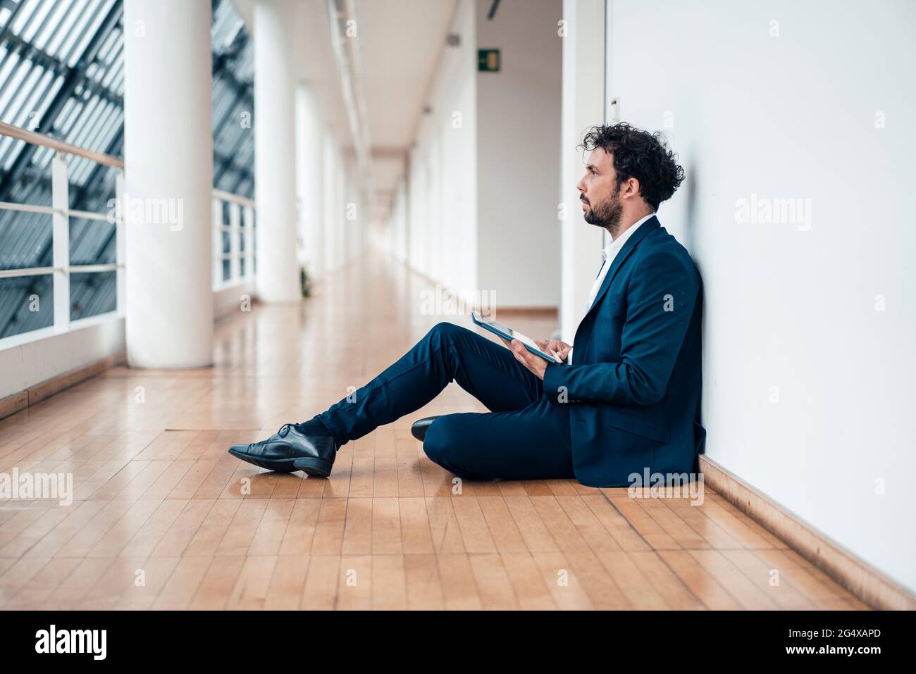 Geschäftsmann mit digitalem Tablet, der auf dem Büroflur sitzt und wegschaut Stockfoto