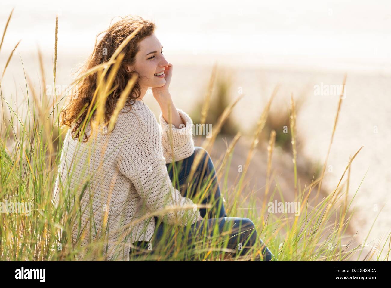 Junge Frau mit der Hand am Kinn, die während des Urlaubs in den Dünen am Strand sitzt Stockfoto