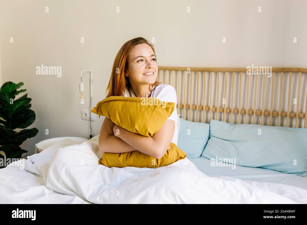 Lächelnde junge Frau, die zu Hause Kissen auf dem Bett umarmt Stockfoto