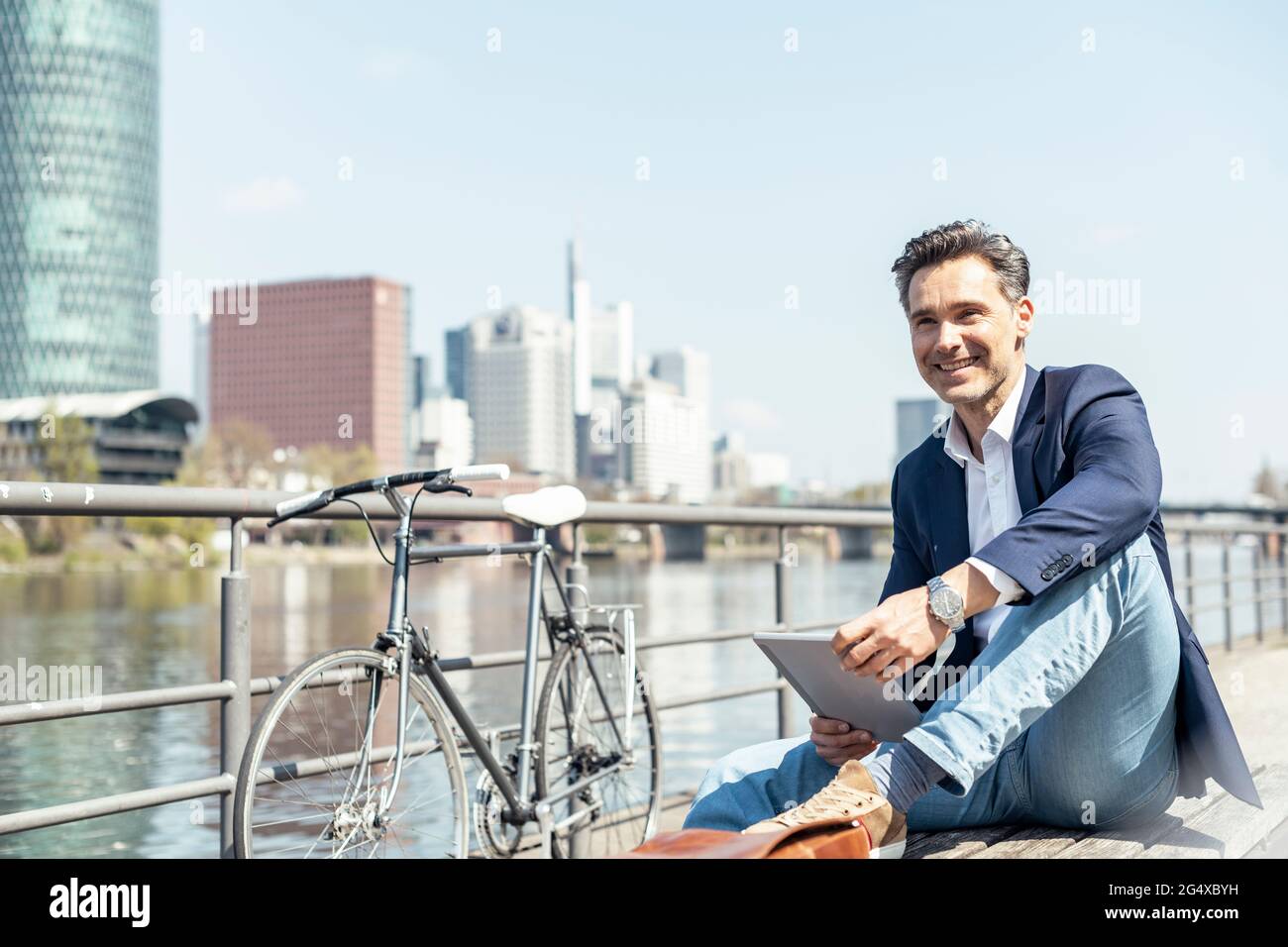 Lächelnder männlicher Unternehmer mit digitalem Tablet, der mit dem Fahrrad in der Stadt sitzt Stockfoto