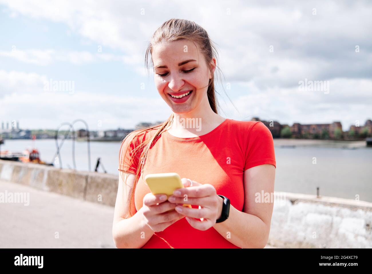 Lächelnde, flaue Frau, SMS-Nachrichten auf dem Smartphone vor dem Himmel Stockfoto