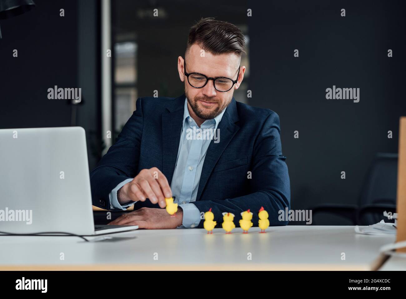 Männlicher Unternehmer mit Laptop, der Baby-Hühnchen-Spielzeug im Büro hält Stockfoto
