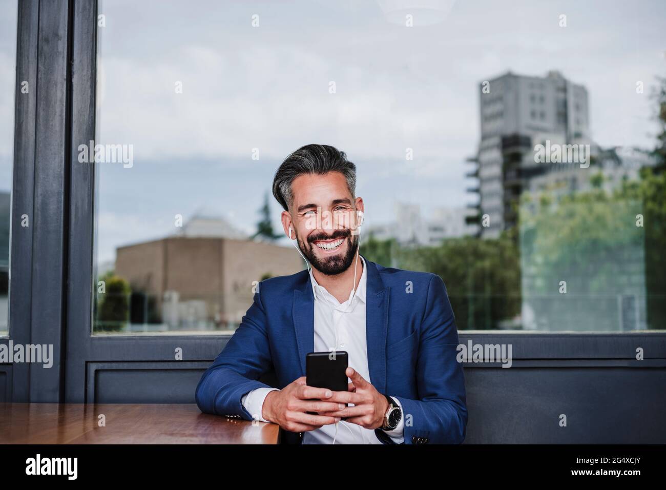 Lächelnder Geschäftsmann mit Ohrhörern und Mobiltelefon, das sich am Fenster auflehnt Stockfoto