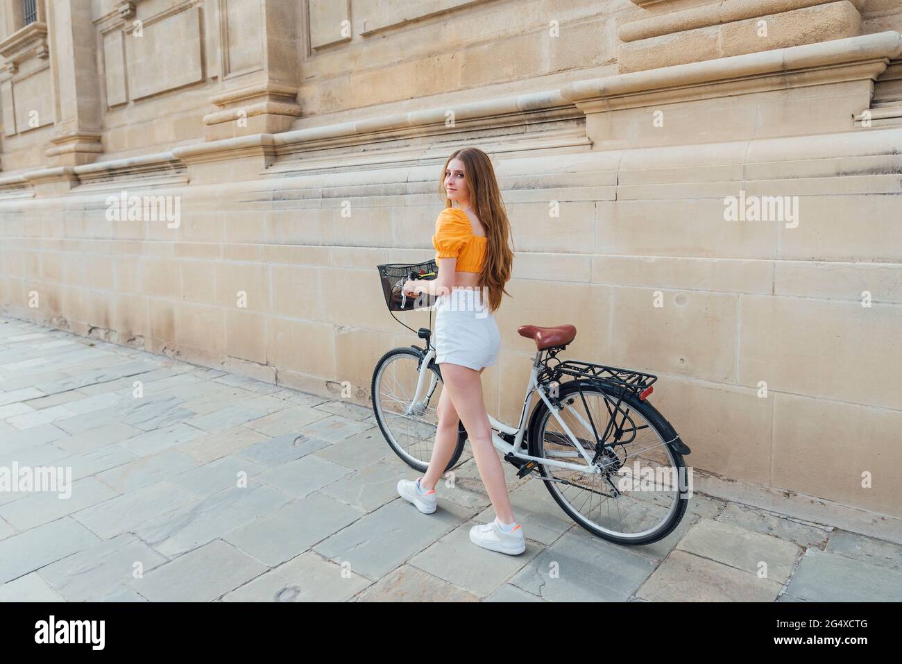 Junge Frau schaut weg, während sie mit dem Fahrrad an einer Steinmauer fährt Stockfoto