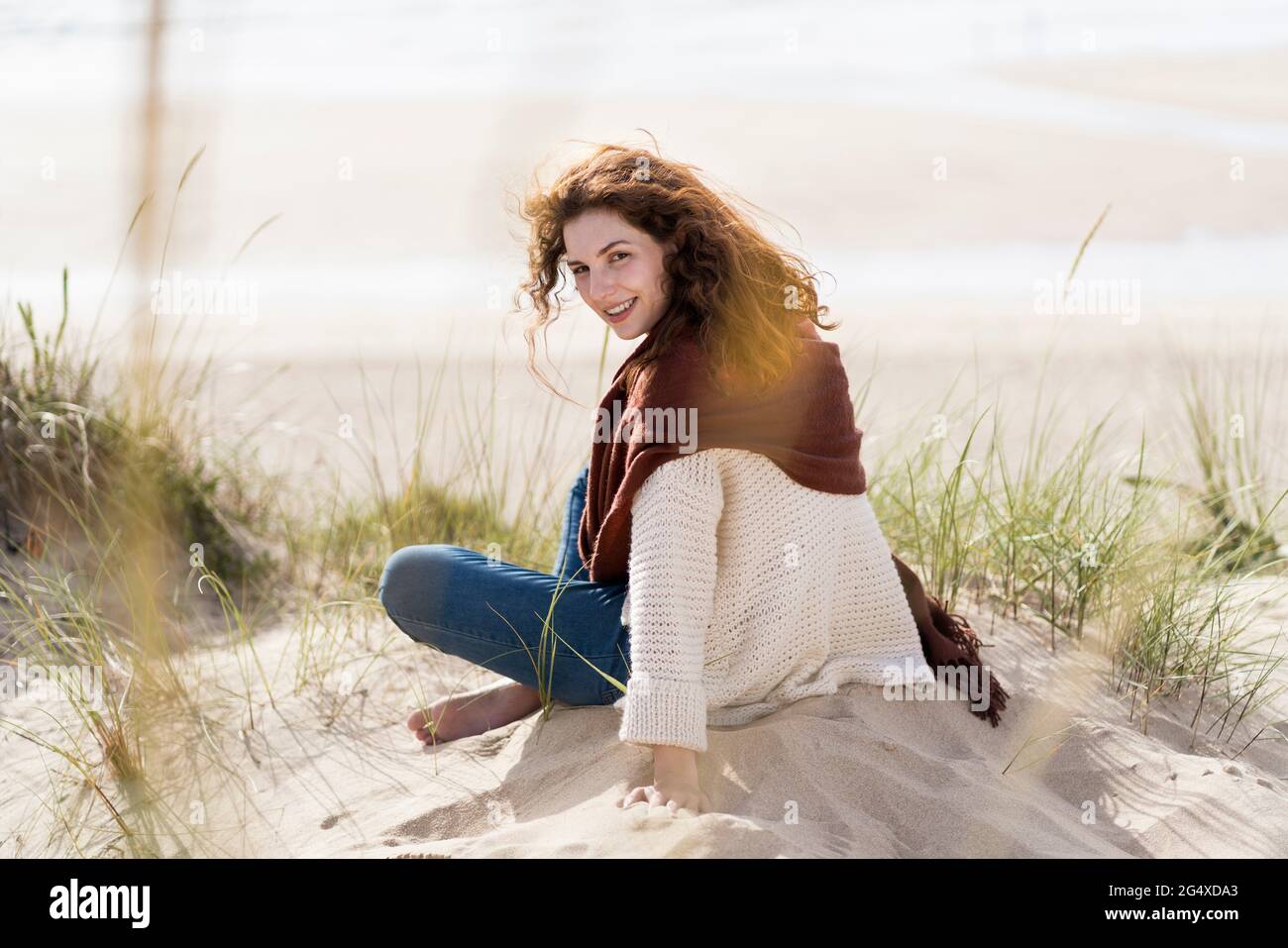 Lächelnde Frau, die über die Schulter blickt, während sie mit gekreuzten Beinen auf Sand am Strand sitzt Stockfoto