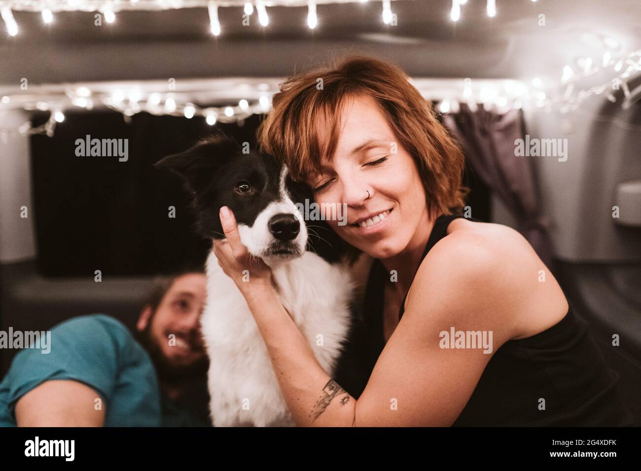 Lächelnde Frau umarmt Hund, während sie in beleuchtetem Van in der Nacht reist Stockfoto