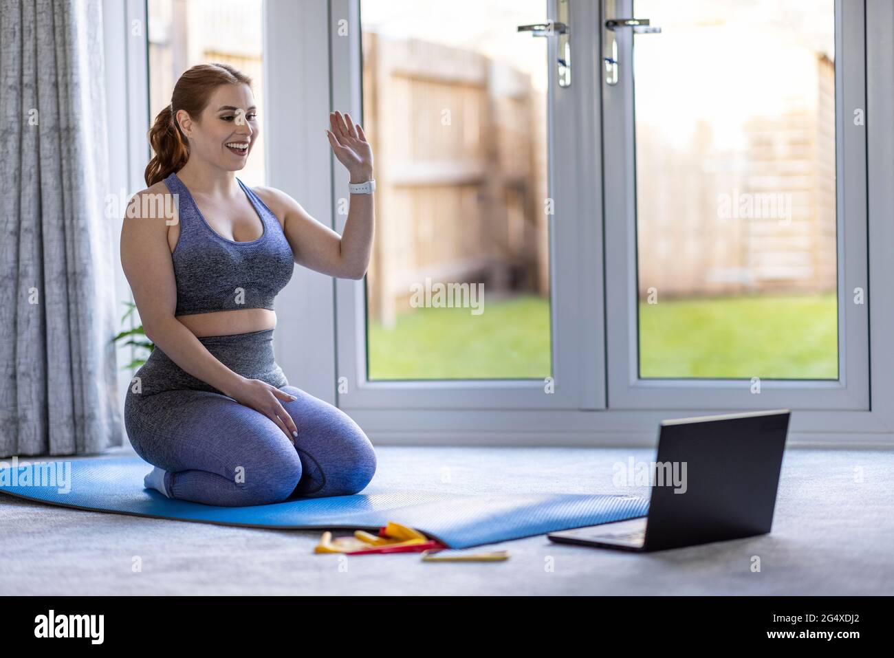 Lächelnde Frau winkt auf dem Laptop, während sie auf der Trainingsmatte sitzt Stockfoto
