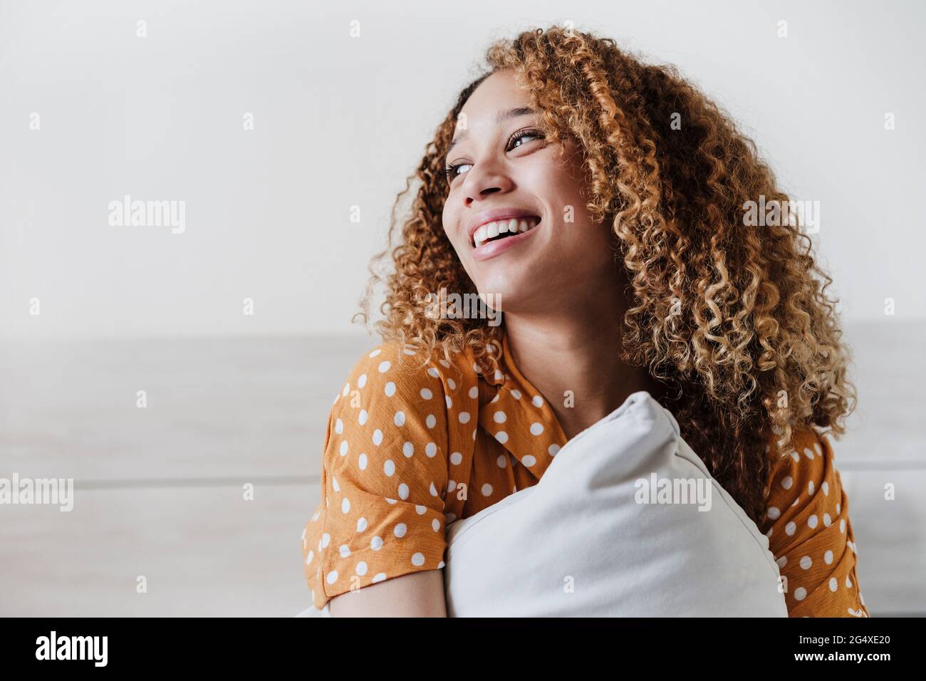Lächelnde, lockige Frau, die zu Hause mit Kissen im Schlafzimmer sitzt Stockfoto