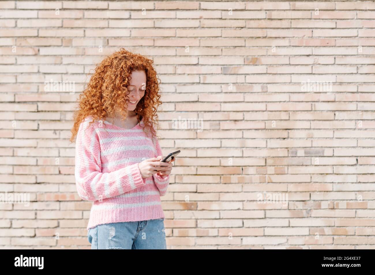 Junge Frau mit lockigen Haaren mit Mobiltelefon, während sie vor einer Ziegelwand steht Stockfoto