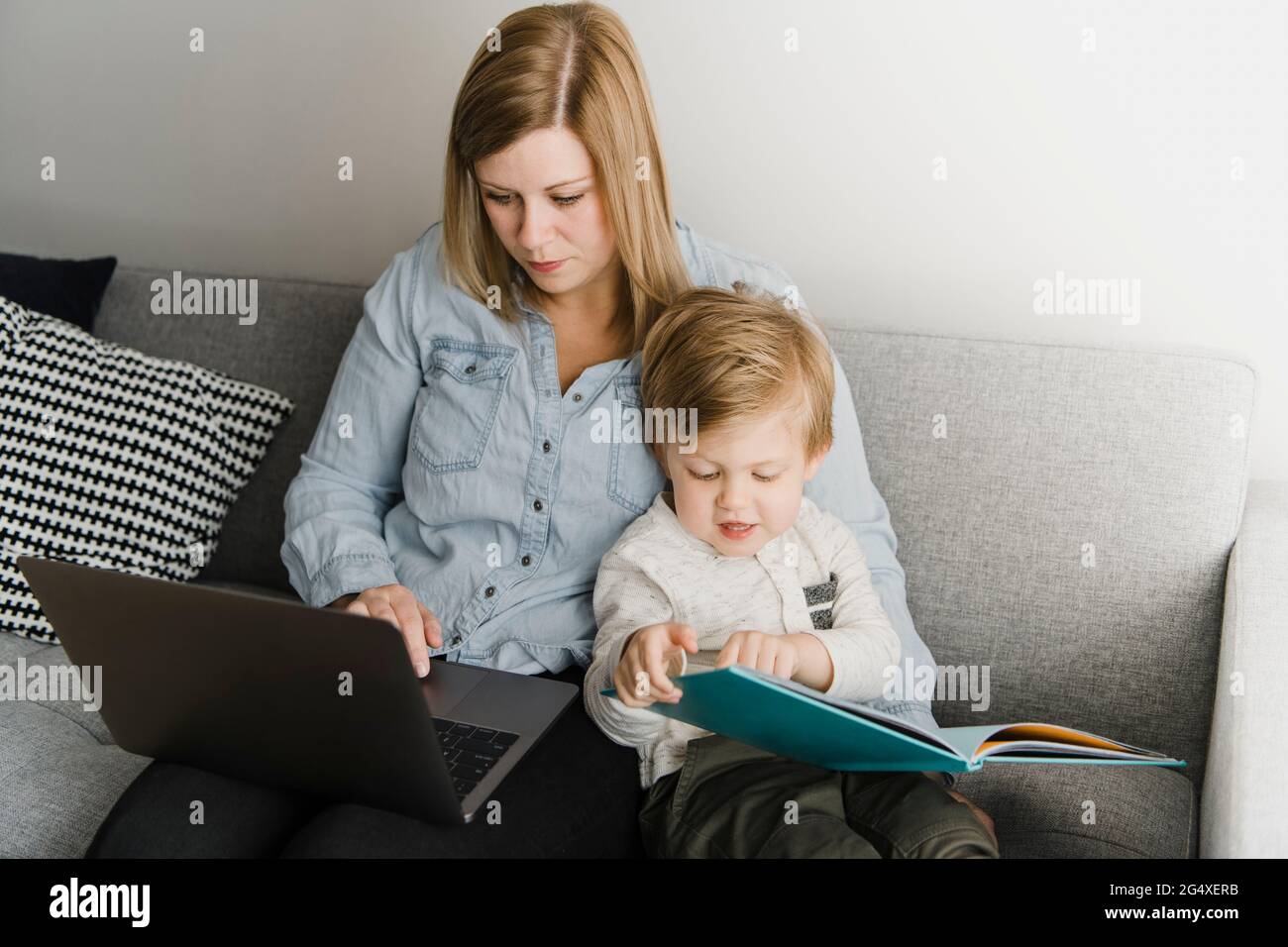 Mutter mit Laptop, während Sohn zu Hause Bilderbuch auf dem Sofa liest Stockfoto