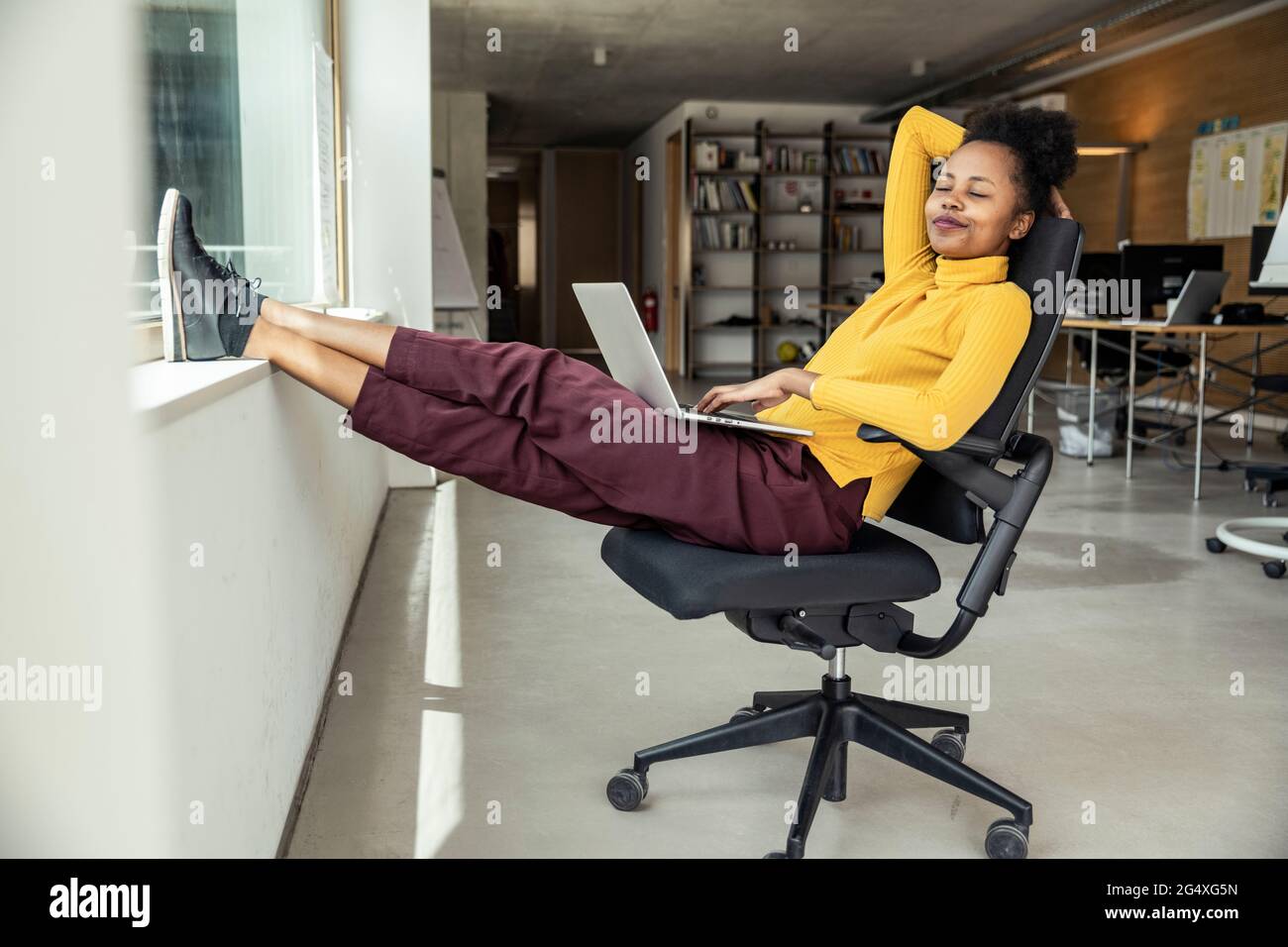 Geschäftsfrau mit Laptop, die sich auf einem Stuhl in der Bürowohnung entspannt Stockfoto