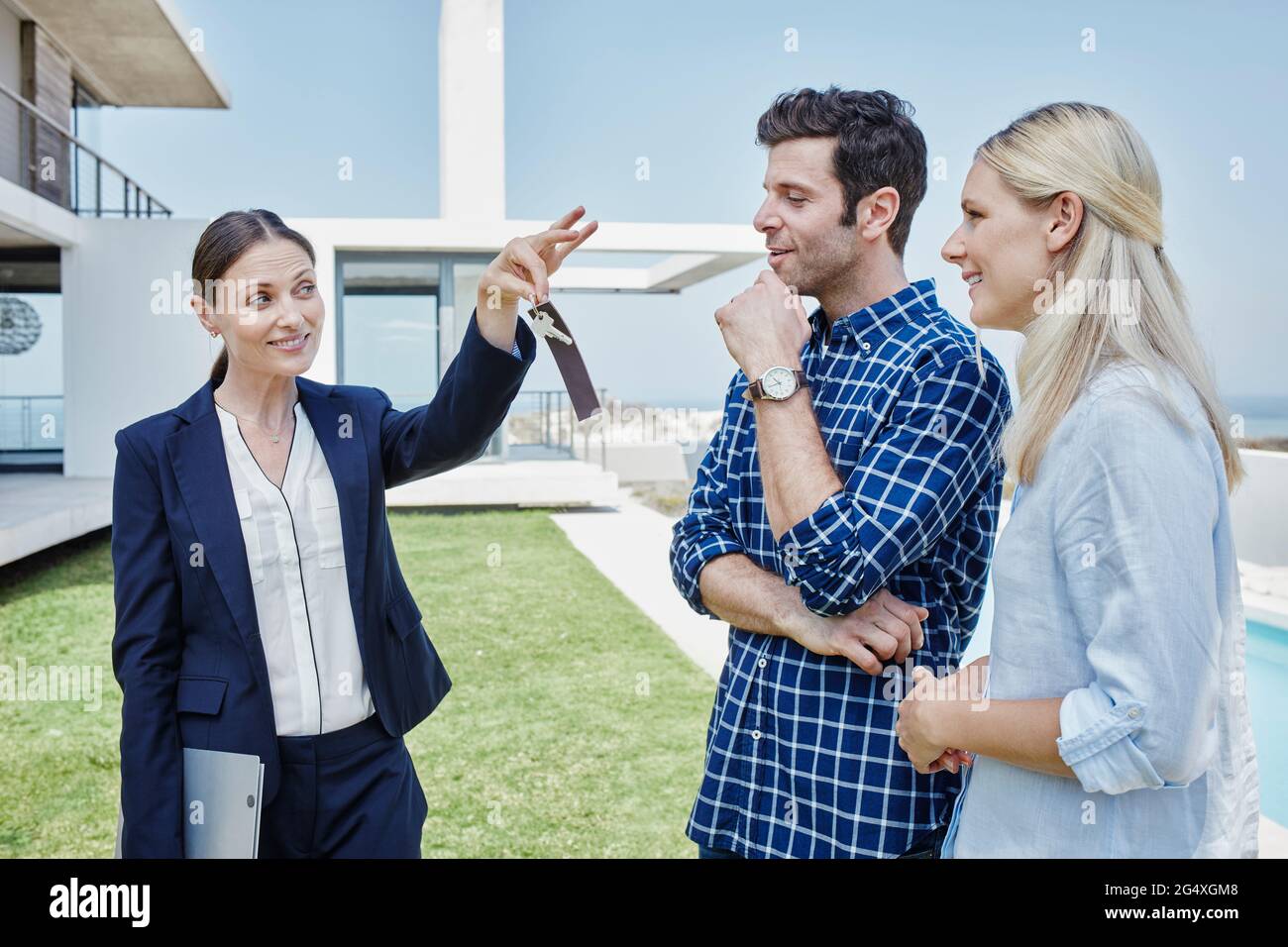 Weibliche Immobilienmaklerin zeigt dem Paar den Hausschlüssel, während sie auf dem Rasen steht Stockfoto