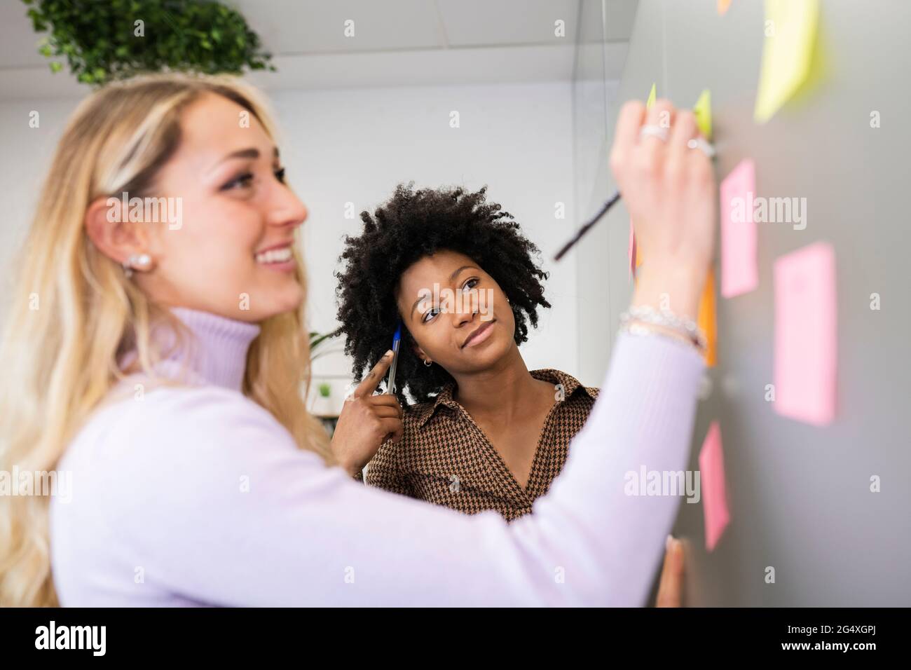 Eine Frau, die beim Schreiben von Haftnotizen auf eine Mitarbeiterin schaut, während sie im Büro eine Strategie plant Stockfoto Eine Frau, die beim Schreiben von Haftnotizen auf eine Mitarbeiterin schaut, während sie im Büro eine Strategie plant Stockfoto